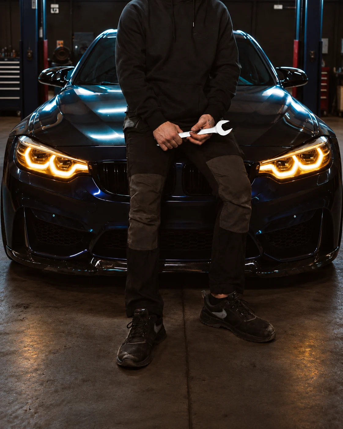 Mehanik s ključem pred vozilom BMW s prižganimi žarometi v delavnici CO. Performance